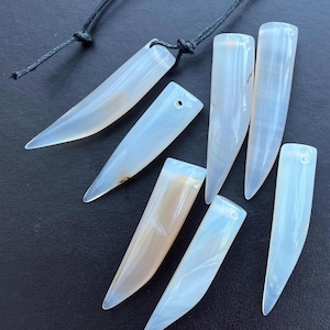 May include: A selection of light blue and white agate pendants, each with a pointed shape and a drilled hole for stringing. Some pendants have a tan hue. A black cord is attached to two of the pendants. The pendants are arranged on a dark surface.
