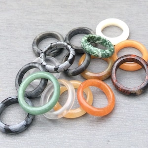 May include: A selection of gemstone rings in various colours and patterns. The rings include shades of green, orange, white, and grey, with speckled and marbled designs. The rings are made of different materials, including jade and other stones.