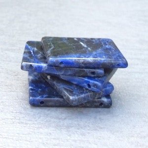 Op de afbeelding: Een stapel van zes vierkante lapis lazuli kralen met twee gaten geboord door elke kraal. De kralen zijn diepblauw met witte en zwarte marmering.