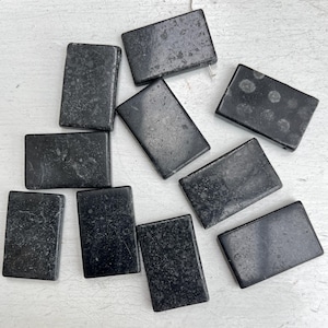 May include: A selection of rectangular, dark grey stone beads with a slightly textured surface. The beads are arranged on a white surface, highlighting their shape and colour variations. Some beads have a speckled appearance.