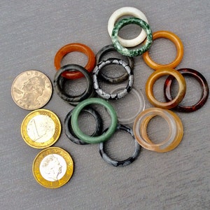 Könnte beinhalten: Eine Sammlung verschiedener Edelsteinringe in Orange-, Grün-, Schwarz- und Klartönen. Ein amerikanischer Vierteldollar und zwei Euro-Münzen dienen als Größenvergleich. Die Ringe variieren in Farbe und Design und zeigen eine Reihe natürlicher Materialien.