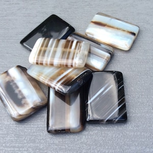 Può includere: Una collezione di otto perline rettangolari in agata in tonalità di marrone, nero e bianco. Le perline hanno un motivo a strisce.