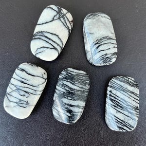 Puede incluir: Cinco piedras rectangulares con bordes redondeados. Las piedras son blancas con patrones negros, similares a vetas, que recorren sus superficies. Están dispuestas sobre una superficie oscura, resaltando sus texturas y colores naturales.