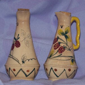 Op de afbeelding: Twee keramische flessen voor olie en azijn met een aardbeidesontwerp. De flessen zijn crèmekleurig met groene bladeren en rode aardbeien. De fles links heeft een kurkstop en de fles rechts heeft een gele handgreep.