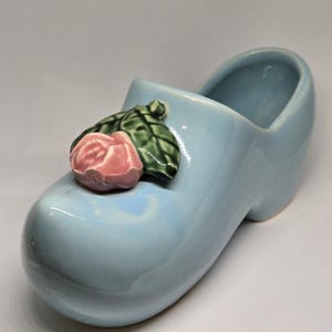 Pode incluir: Um vaso de cerâmica azul claro em forma de tamanco, adornado com uma rosa rosa e folhas verdes. O vaso decorativo tem um acabamento brilhante e é provavelmente para uso interno.