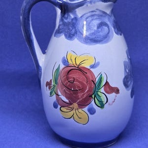 May include: White ceramic pitcher with a blue handle and rim, decorated with a red rose and yellow and green floral accents. The pitcher has a curved handle and a rounded body, suitable for serving beverages.