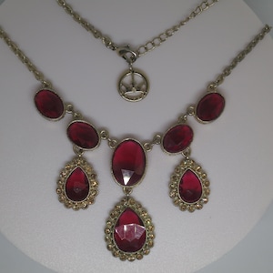 Op de afbeelding: Een gouden ketting met een kroonhanger en een rode strass-statementstuk. Het statementstuk heeft een grote, druppelvormige strass in het midden, omringd door kleinere strasssteentjes.