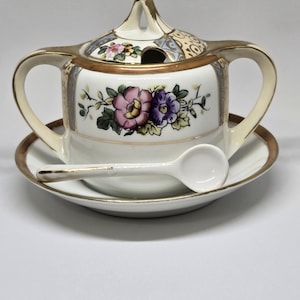 Puede incluir: Un azucarero de porcelana vintage con tapa, dos asas y un platillo a juego. El cuenco está decorado con diseños florales en rosa y morado, con ribetes dorados. Una pequeña cuchara blanca descansa sobre el platillo.