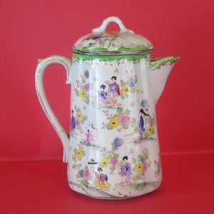 Può includere: Una teiera in porcellana bianca con un coperchio verde e disegni floreali e di geisha. La teiera ha un manico e un beccuccio.