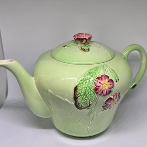 May include: A light green ceramic teapot with a matching lid. The teapot features a floral design with pink flowers and green leaves. The handle and spout are curved. The lid has a small pink flower as a handle.