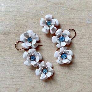 Op de afbeelding: Een paar oorbellen met elk drie witte bloemencharms. De bloemen hebben een blauwe strasssteen in het midden.