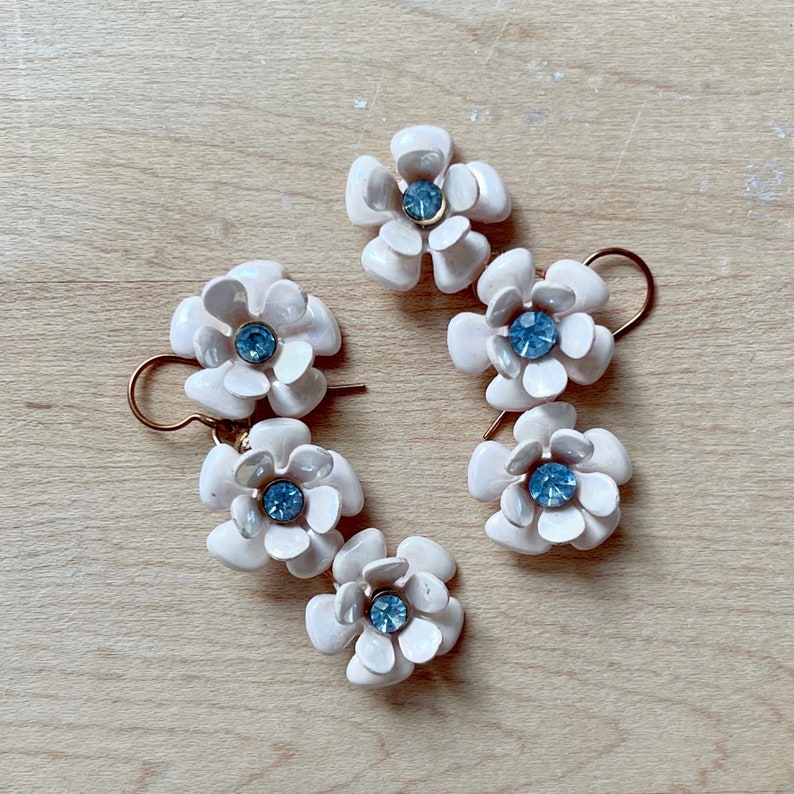 Op de afbeelding: Een paar oorbellen met elk drie witte bloemencharms. De bloemen hebben een blauwe strasssteen in het midden.