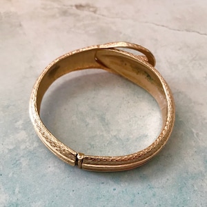 May include: A gold-toned hinged bangle bracelet with a textured finish. The bracelet has a unique design with a curved, overlapping section.