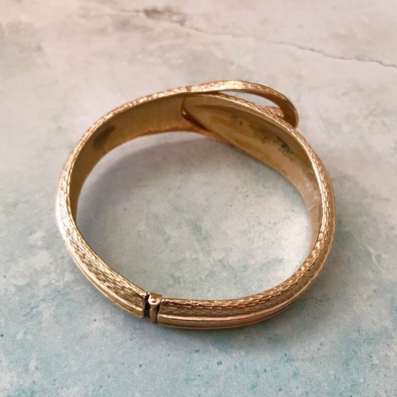 May include: A gold-toned hinged bangle bracelet with a textured finish. The bracelet has a unique design with a curved, overlapping section.