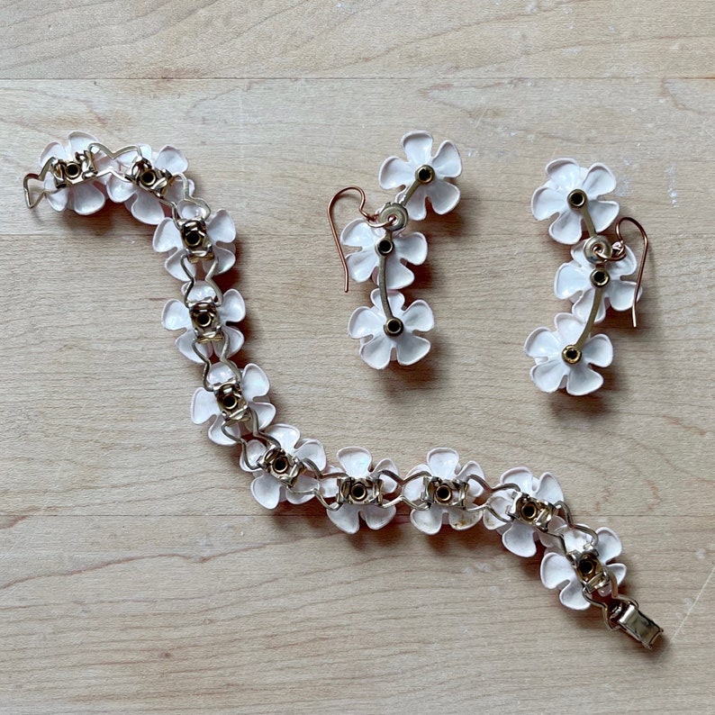 Op de afbeelding: Een witte, bloemvormige armband en bijpassende oorbellen. De sieraden zijn gemaakt van metaal met witte emaille bloemen en gouden accenten.