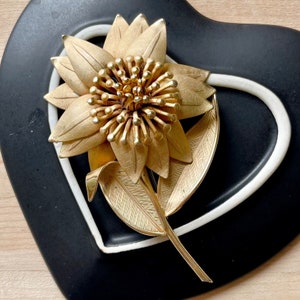 May include: A gold-toned flower brooch with a detailed design. The flower has multiple petals and a center with small, round accents. The brooch is set against a black and white background.