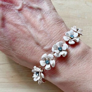 Op de afbeelding: Een witte bloemenarmband met blauwe strass accenten. De armband is gemaakt van metaal en heeft een delicaat kettingontwerp.