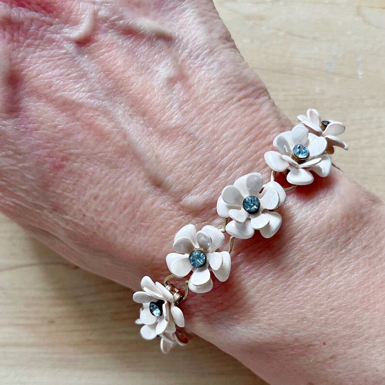 Op de afbeelding: Een witte bloemenarmband met blauwe strass accenten. De armband is gemaakt van metaal en heeft een delicaat kettingontwerp.