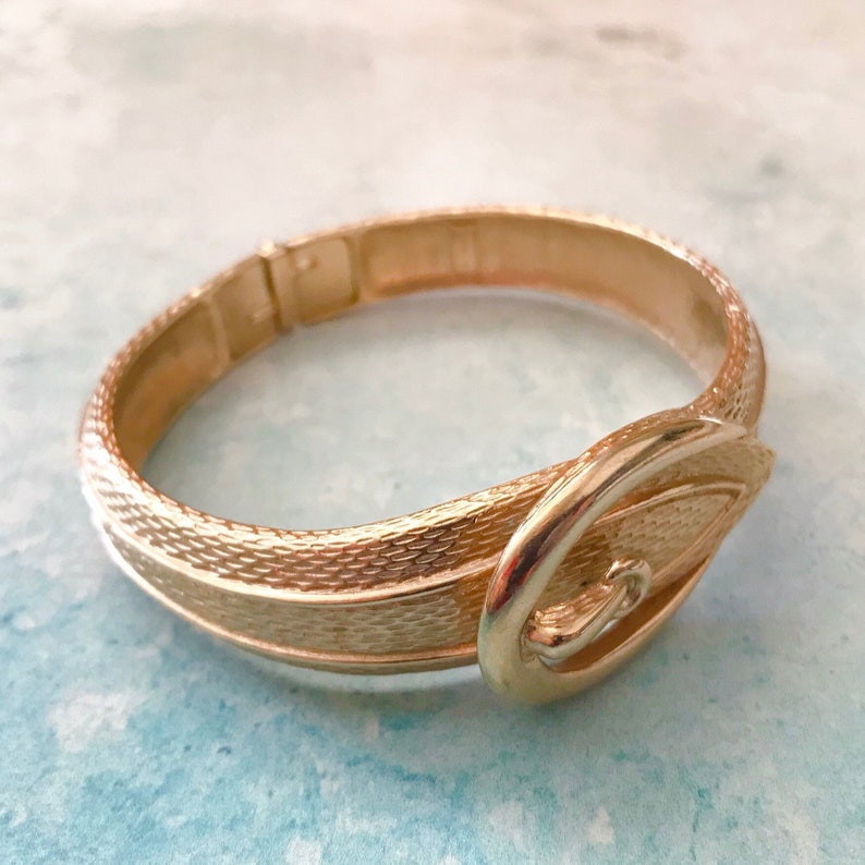 May include: A gold-toned metal bracelet with a buckle design. The bracelet is textured and has a shiny finish.