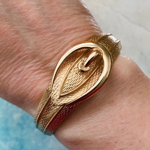 May include: A gold-toned metal bracelet with a large, textured buckle in the shape of a teardrop.