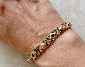 Puede incluir: Un brazalete de oro con esmalte negro y diamantes de imitación transparentes. El brazalete está hecho de formas geométricas enlazadas.