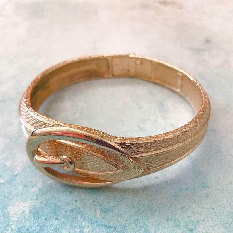 May include: A gold-tone metal bangle bracelet with a textured finish and a buckle design.