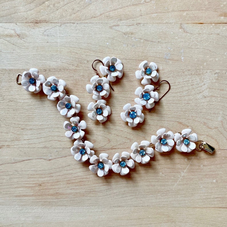 Op de afbeelding: Een witte keramische bloemenarmband en bijpassende oorbellen met blauwe strass steentjes in het midden. De sieraden liggen op een licht houten oppervlak.
