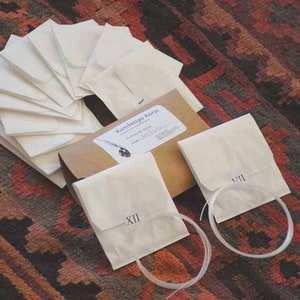 May include: A brown paper bag with the text "Kumbengo Kord" and a drawing of a string instrument. The bag is filled with white paper envelopes, some of which have Roman numerals written on them.