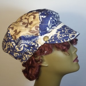 May include: A blue, beige, and brown patterned fabric newsboy cap with a white trim and a gold button. The cap is quilted and has a slightly rounded crown.