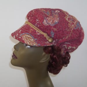 May include: A quilted, maroon newsboy cap with a floral pattern in shades of beige, blue, and gold. The cap features a small brim, a decorative rope detail, and a gold button. The hat is a stylish accessory.