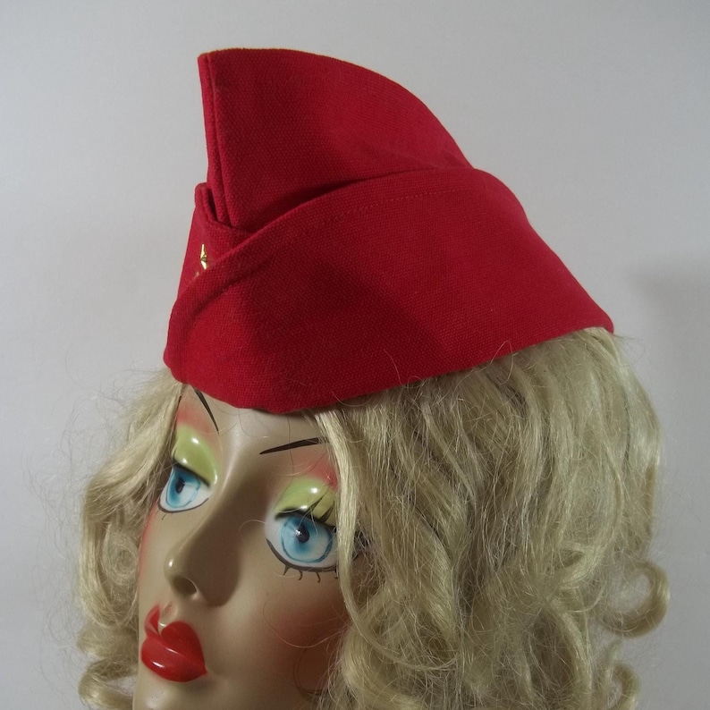 May include: A bright red vintage-style hat with a folded brim and a small, decorative gold star. The hat is made of a felt-like material and has a classic, military-inspired design. The hat is a striking accessory.