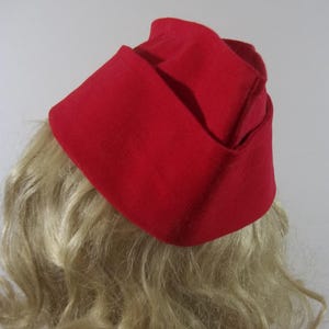May include: A bright red hat with a folded design. The hat is made of a solid, textured fabric and sits atop wavy blonde hair. The hat's shape is reminiscent of a vintage style, with a folded brim and a structured crown.