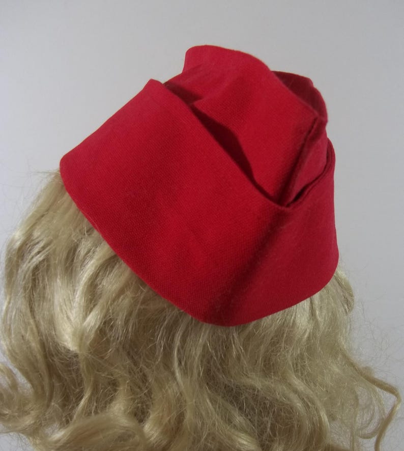 May include: A bright red hat with a folded design. The hat is made of a solid, textured fabric and sits atop wavy blonde hair. The hat's shape is reminiscent of a vintage style, with a folded brim and a structured crown.