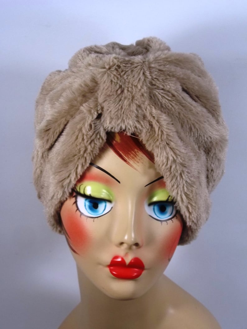 May include: A beige faux fur turban hat with a unique folded design. The hat is soft and fluffy, and it would be a stylish addition to any winter wardrobe.
