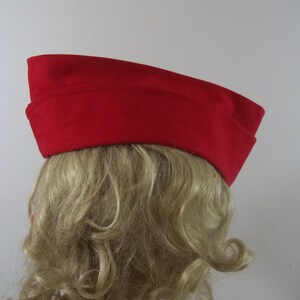 May include: A bright red hat with a rectangular crown and a slightly curved brim. The hat is made of a felt-like material and is worn on a person's head. The hat is a solid color and has a simple, classic design.