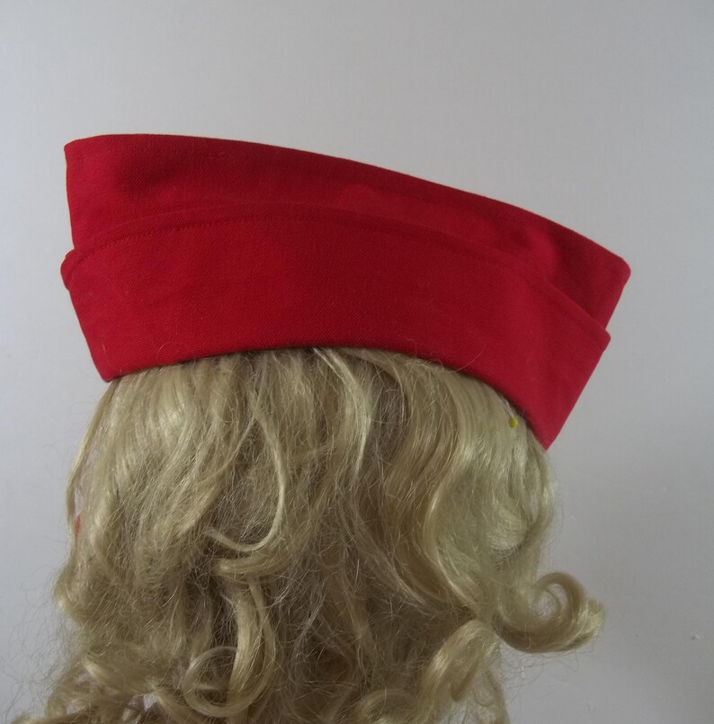 May include: A bright red hat with a rectangular crown and a slightly curved brim. The hat is made of a felt-like material and is worn on a person's head. The hat is a solid color and has a simple, classic design.