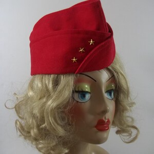 May include: A red felt hat with three gold stars. The hat has a folded design and is worn on a mannequin head with blonde hair, blue eyes, and red lipstick. The hat is a vintage style.