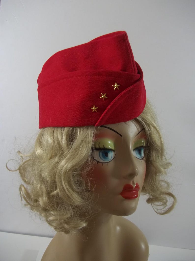 May include: A red felt hat with three gold stars. The hat has a folded design and is worn on a mannequin head with blonde hair, blue eyes, and red lipstick. The hat is a vintage style.