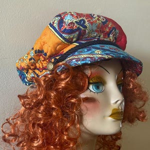 May include: A colorful, patterned fabric hat with a wide brim. The hat is made of a patchwork of different fabrics, including orange, blue, red, and yellow. The hat is worn by a mannequin with red curly hair.