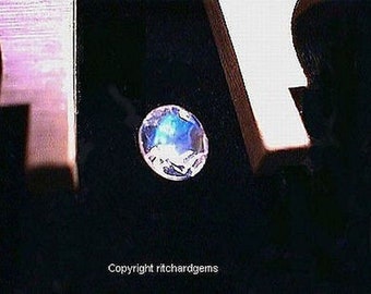 May include: A close-up of a round, iridescent gemstone with a blue and white color scheme. The gemstone is set against a black background.