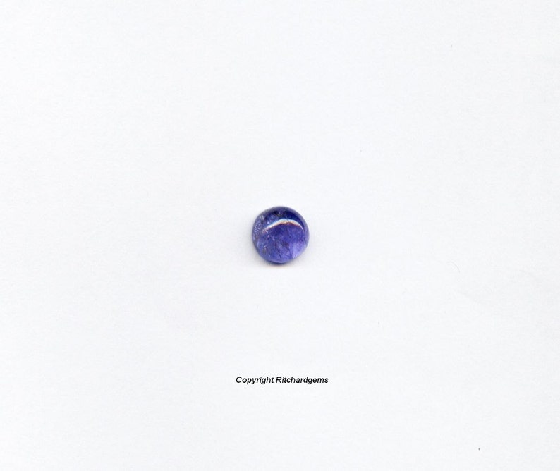 Puede incluir: Una piedra preciosa redonda de color azul con un acabado ligeramente iridiscente. La piedra preciosa se coloca sobre un fondo blanco.