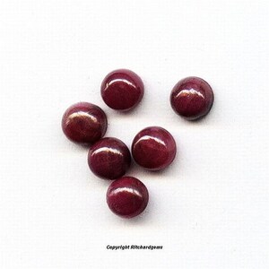 May include: Seven round, dark red gemstones, likely rubies, are scattered on a white surface.
