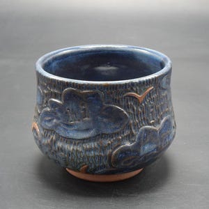 Könnte beinhalten: Eine blaue Keramikschüssel mit einer strukturierten Oberfläche und einem Design aus Wolken und Vögeln. Die Schüssel hat einen leicht abgerundeten Boden und eine breite Öffnung.