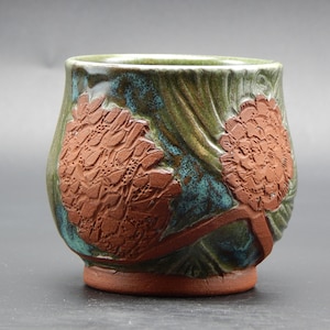 Może przedstawiać: Zielony kubek ceramiczny z brązową glazurą i dwoma wypukłymi wzorami kwiatowymi.
