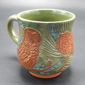 Może przedstawiać: Zielony kubek ceramiczny z wzorem brązowej sowy i szyszek. Sowa znajduje się w środku kubka, a szyszki wokół niej.