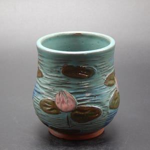 May include: A handcrafted ceramic tumbler with a turquoise glaze. The exterior features a carved design of lily pads and a pink lotus flower. The base is a reddish-brown color. The tumbler is a unique piece of art.