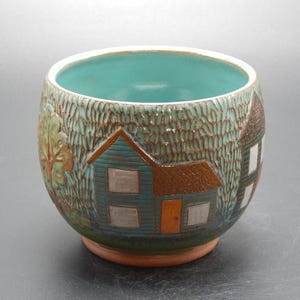 May include: A ceramic bowl with a teal interior and a textured exterior featuring a village scene. The bowl's exterior is a light teal color with brown accents, depicting houses, trees, and foliage. The rim is white, and the base is a reddish-brown.