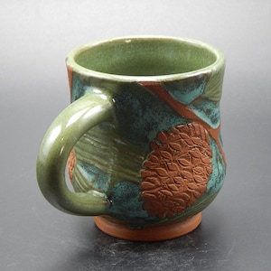 Może przedstawiać: Ręcznie robiony ceramiczny kubek z zielonym uchwytem i wnętrzem. Zewnętrzna strona ma teksturowany wzór z odcieniami zieleni, turkusu i brązu. Kubek ma pojemność około 350 ml.