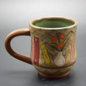 May include: Hand-painted ceramic mug with a brown handle and interior. The exterior features a zigzag pattern, with illustrations of books and a vase of orange flowers. The books are labeled "Blue Skies", "Family", "Bread", and "Long Walks".