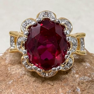 May include: A gold ring featuring a large, oval-cut, deep red gemstone at the center. The stone is surrounded by a scalloped halo of small, clear, sparkling stones set in silver-toned metal. The band is gold with additional small clear stones.
